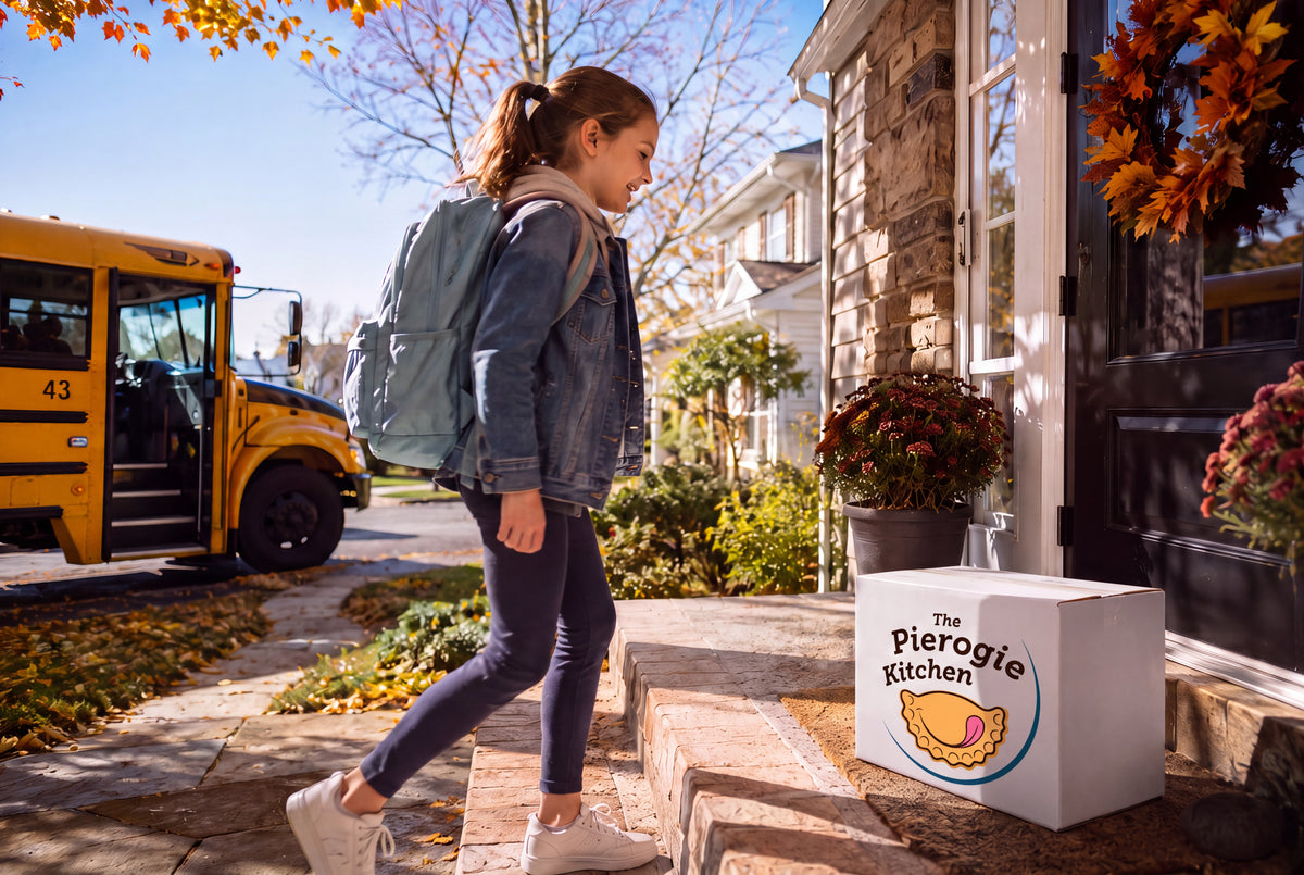 A woman with a gray backpack walks down stone steps toward a house door, with a white delivery box labeled 'The Pierogie Kitchen' on the steps and a yellow school bus parked on the street beside her.
